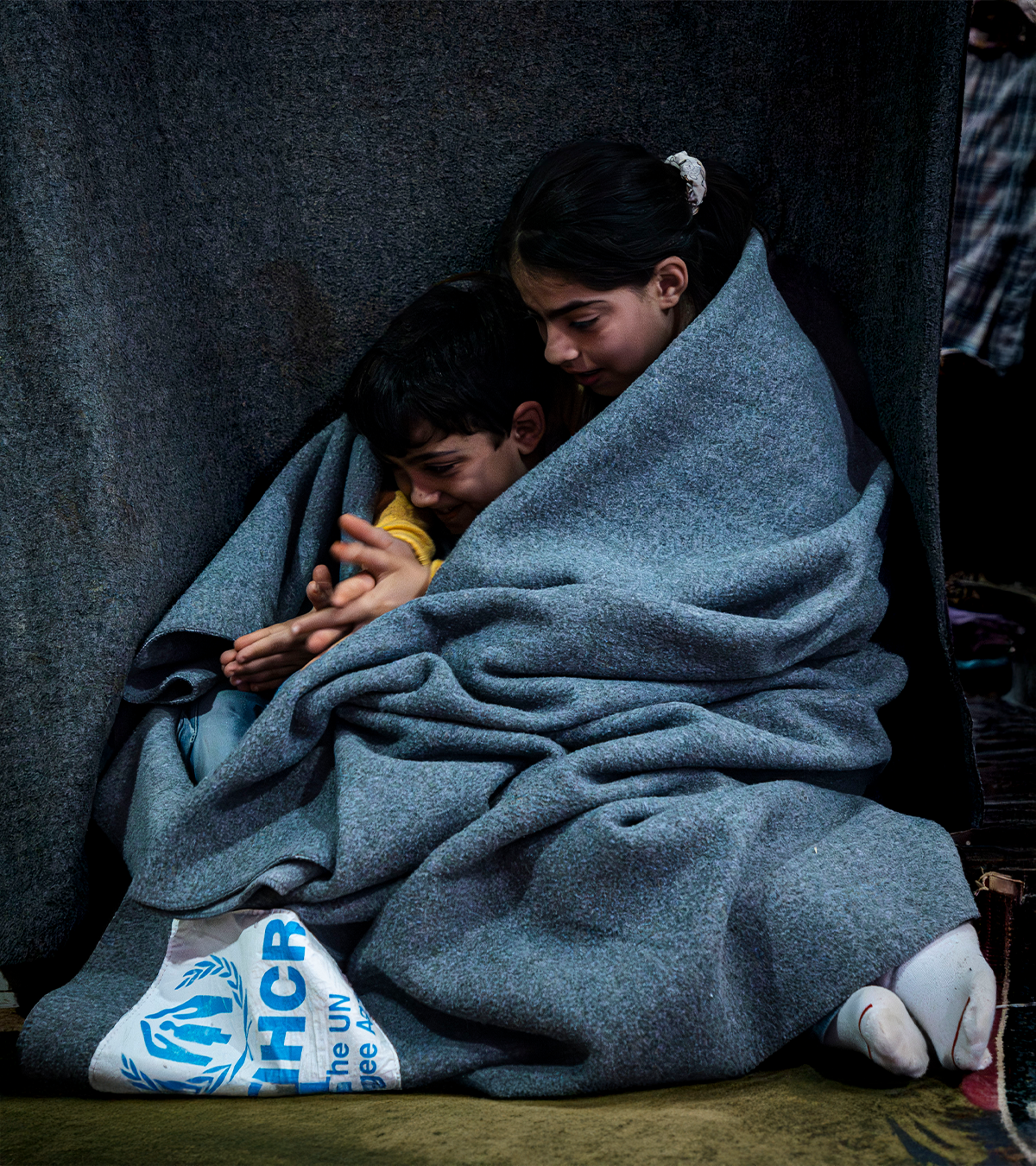 Duas crianças abraçadas envolvidas em um cobertor do ACNUR. Elas estão sorrindo e brincando com as mãos.